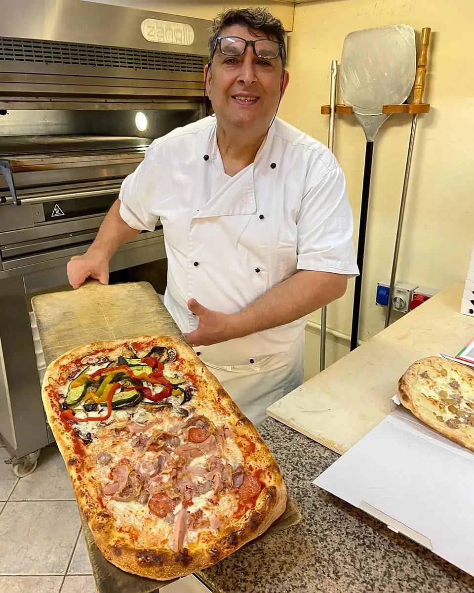 Il pizzaiolo davanti al forno con una pizza al mezzo metro a due gusti, verdure e salumi